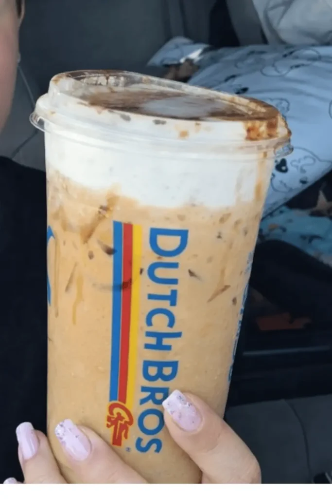 Hand holding a Dutch Bros iced coffee with a caramel-colored base and white foam topping, featuring the Dutch Bros logo stripe on the cup.