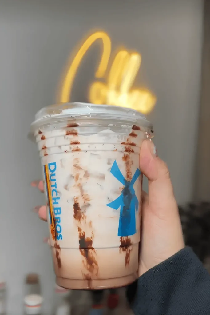 Hand holding a Dutch Bros iced coffee with chocolate drizzle inside the cup and a blue windmill logo, with a blurred "Coffee" neon sign in the background.