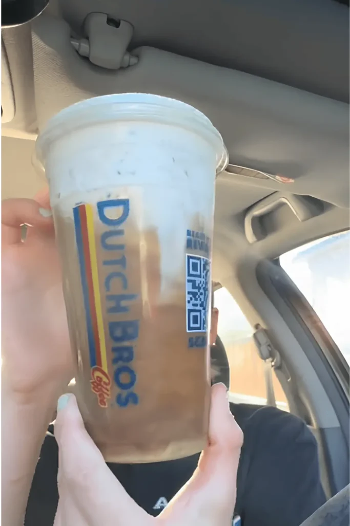 Close-up of a hand holding a Dutch Bros iced coffee with a creamy topping, seen from inside a car, showing the Dutch Bros logo and a QR code on the cup.