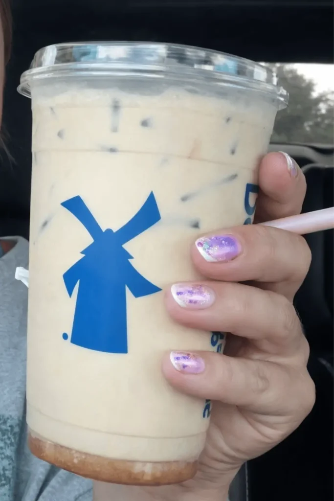 Close-up of a hand with glittery purple and white nails holding a Dutch Bros iced latte with a blue windmill logo on the cup, in a car interior.