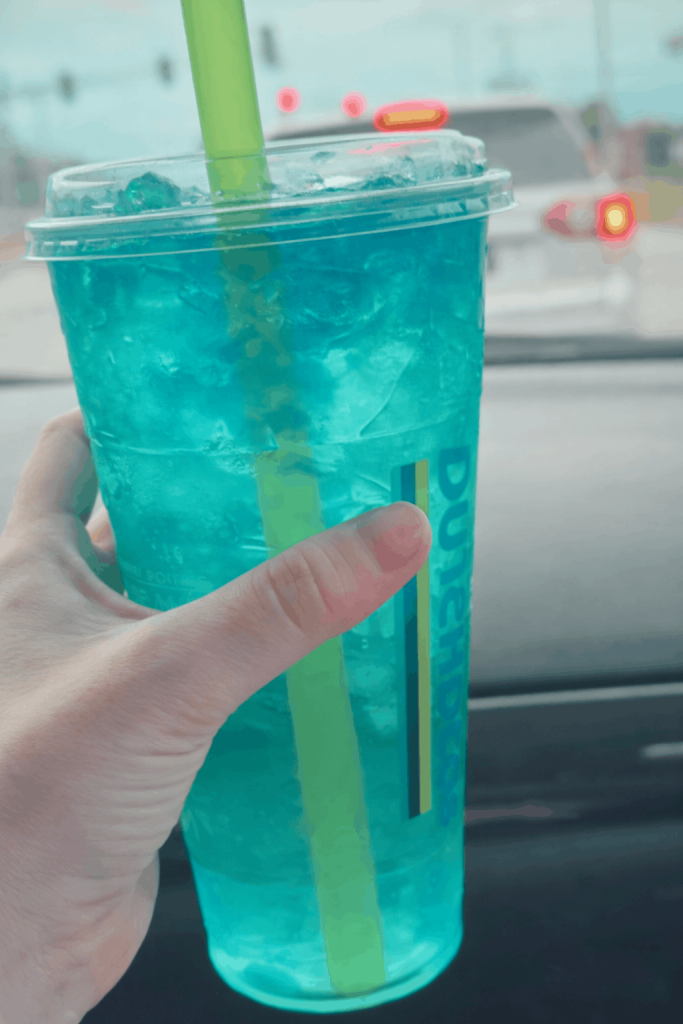A hand holds a clear Dutch Bros cup filled with a bright blue iced drink and a green straw, visible inside a car with traffic lights in the blurred background.