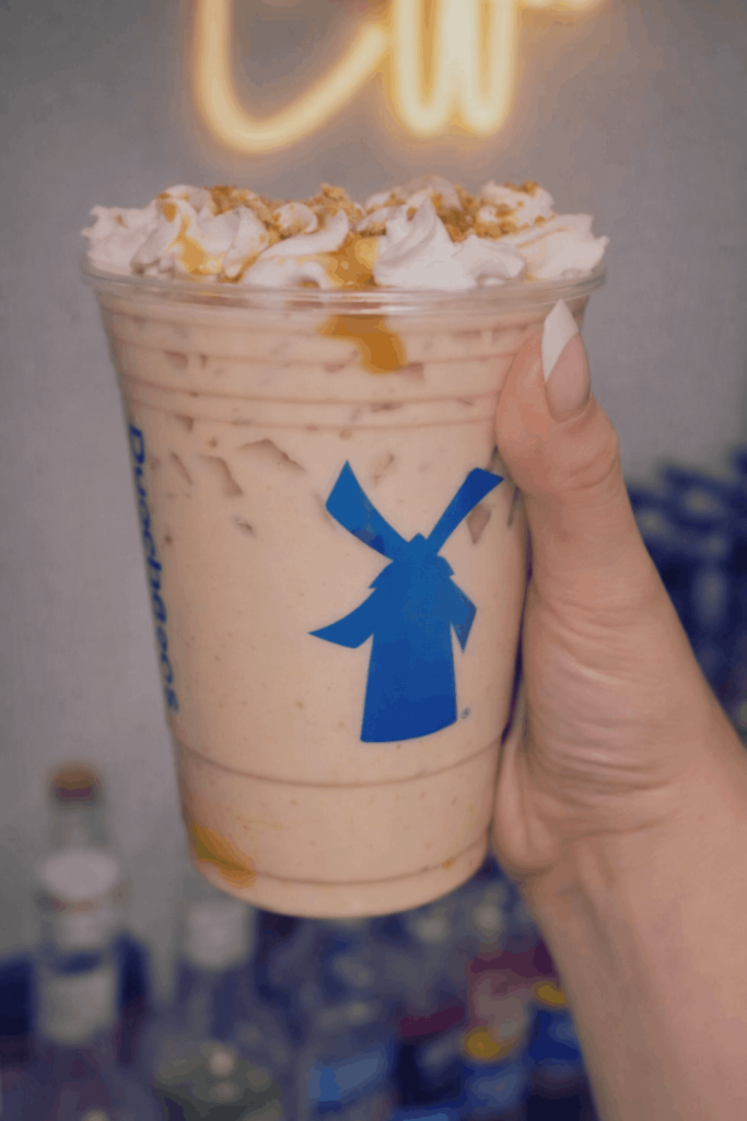 A hand holds a Dutch Bros horchata chai with whipped cream and caramel drizzle, topped with crumbled toppings, against a blurred background with a neon sign.