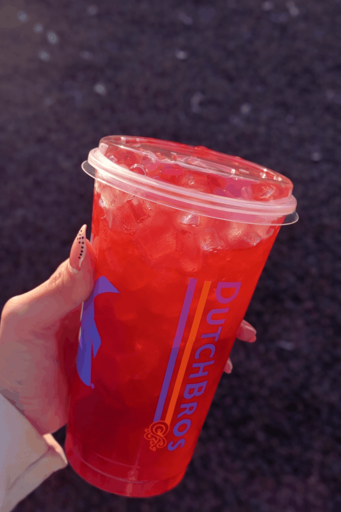 A hand with polka dot nails holds a Dutch Bros cup filled with a vibrant red iced drink, outdoors with green grass in the background.