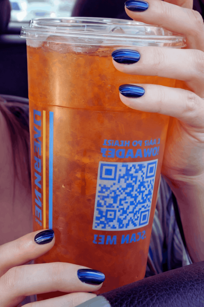 A person's hand with dark blue painted nails holds a Dutch Bros iced tea in a clear cup, showing a blue QR code and "DUTCHBROS" text on the side.