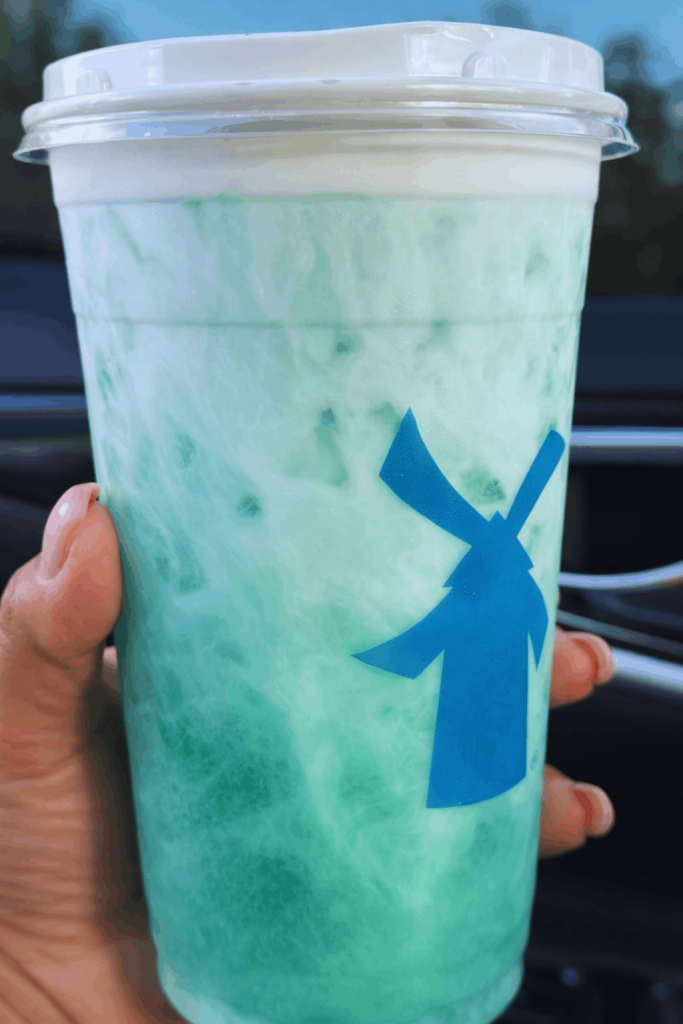 A hand holds a Dutch Bros blended green drink with white swirls and a foamy top, seen from inside a car with a blurred background.