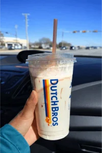Dutch Bros vanilla Shake in a white cup with colorful Dutch Bros logo stripes, held inside a car