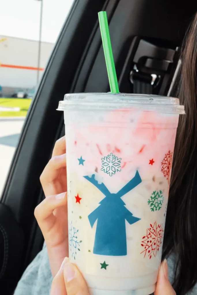 Hand holding a dutch bros Christmas Morning Chai holiday drink in a festive cup with windmill and snowflake design, green straw, inside a car