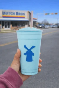 Dutch Bros Cotton Candy Shake in a bright blue frozen cup with blue windmill logo, held in hand outside Dutch Bros store