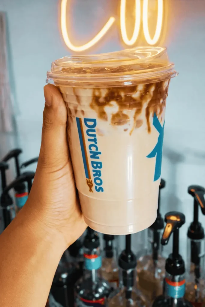 Hand holding a Dutch Bros iced coffee with caramel drizzle in a clear plastic cup, featuring the Dutch Bros logo, with a glowing neon sign in the background