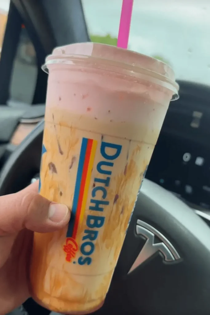  Hand holding a large Dutch Bros iced coffee with a pink cold foam topping, visible inside a Tesla car with the steering wheel and dashboard in the background.