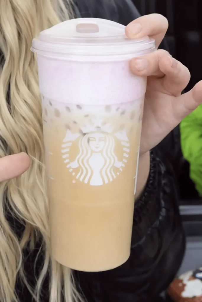 Close-up of a hand holding a Starbucks iced coffee with a prominent pink cold foam topping, showing the Starbucks siren logo on the cup against a blurred background.